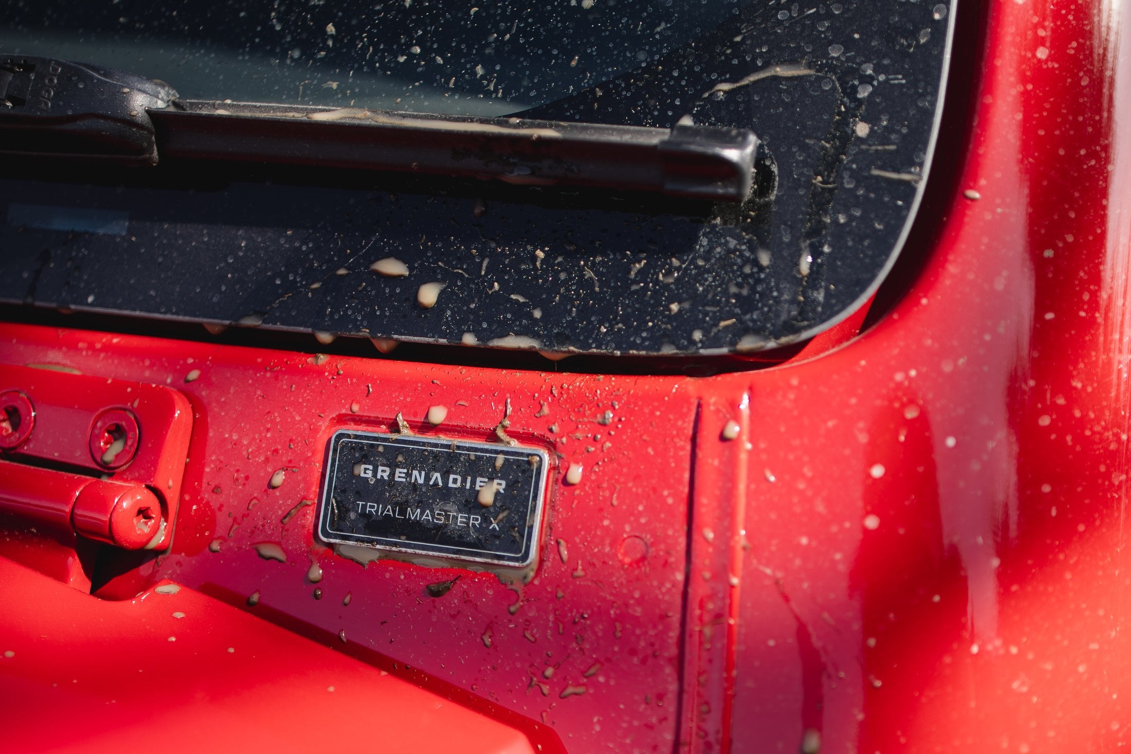 Official close-up of the Grenadier Trialmaster X hood badge on a mud-splattered Devil Red body