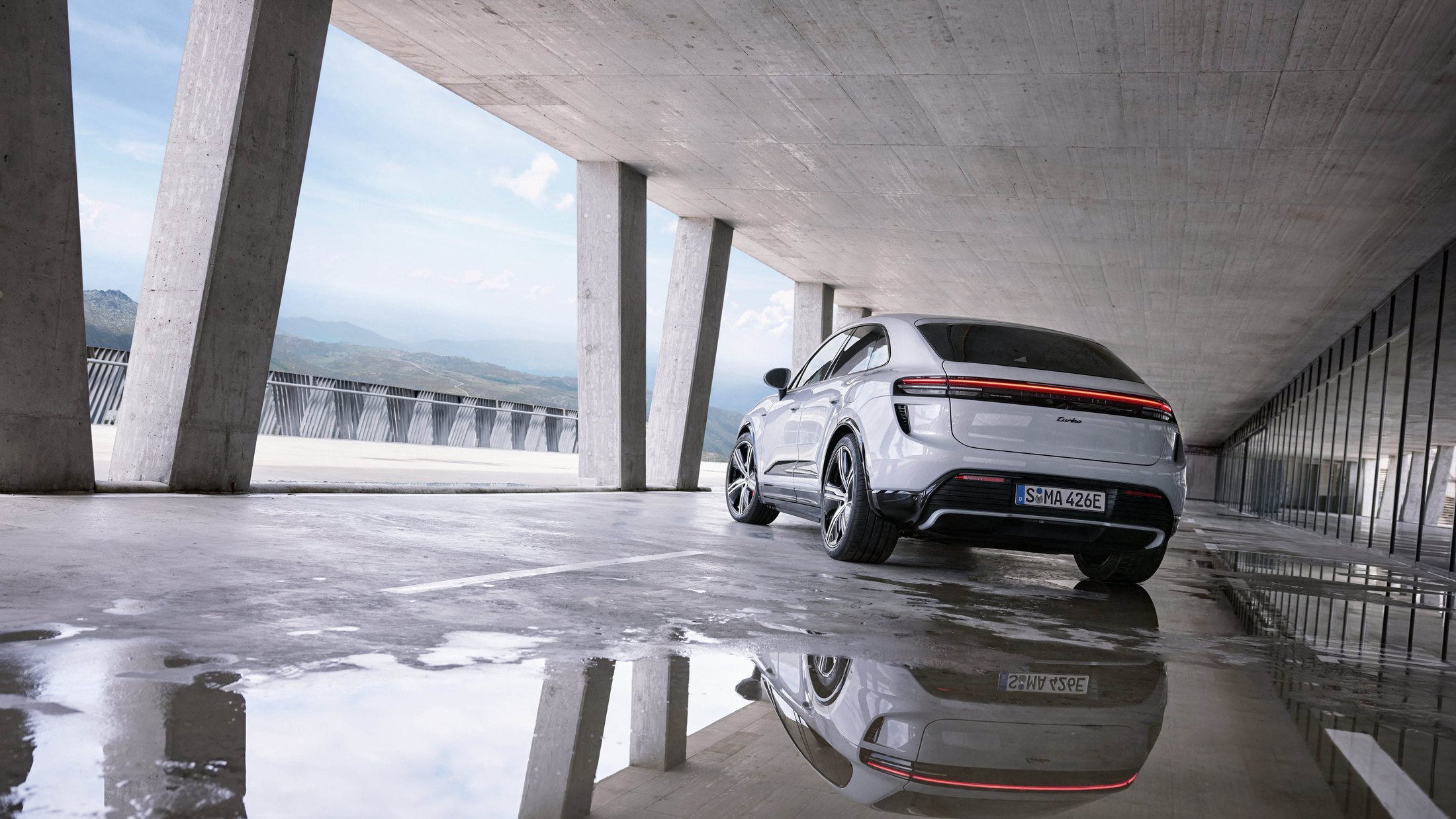 Official Porsche photo of a white Macan Turbo Electric, rear three-quarter in a concrete pavilion