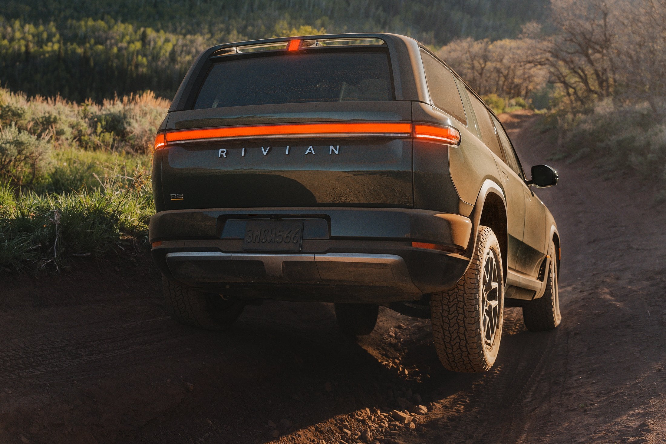 Official Rivian photo of the R2 rear three-quarter on a dirt trail, red light bar lit