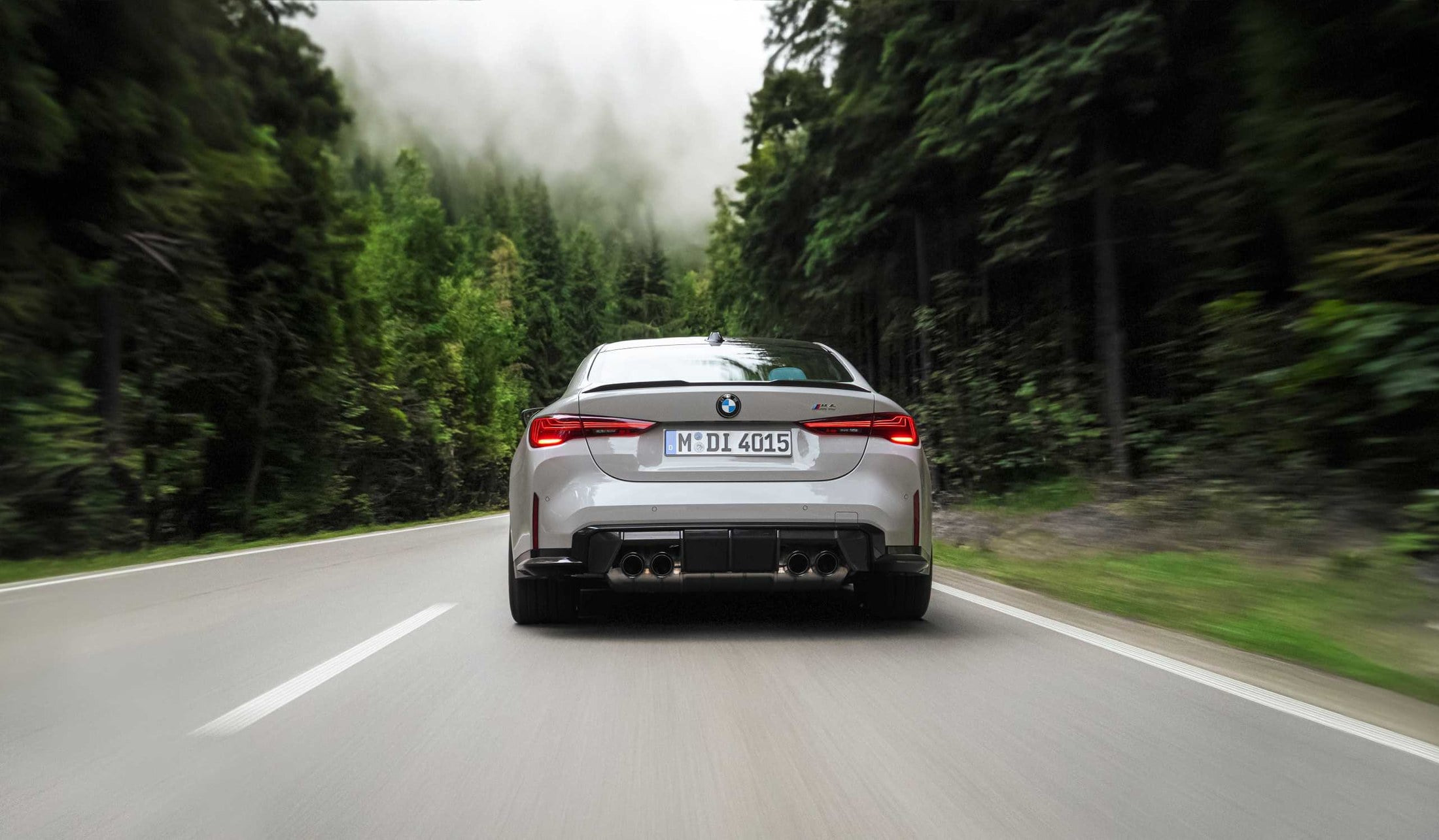 Official BMW photo of a grey M4 Coupe rear three-quarter accelerating away on a forest road