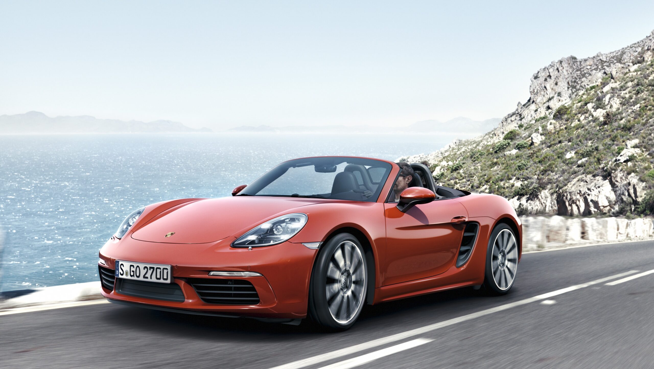 Official Porsche photo of Lava Orange 718 Boxster front three-quarter open-top coastal drive