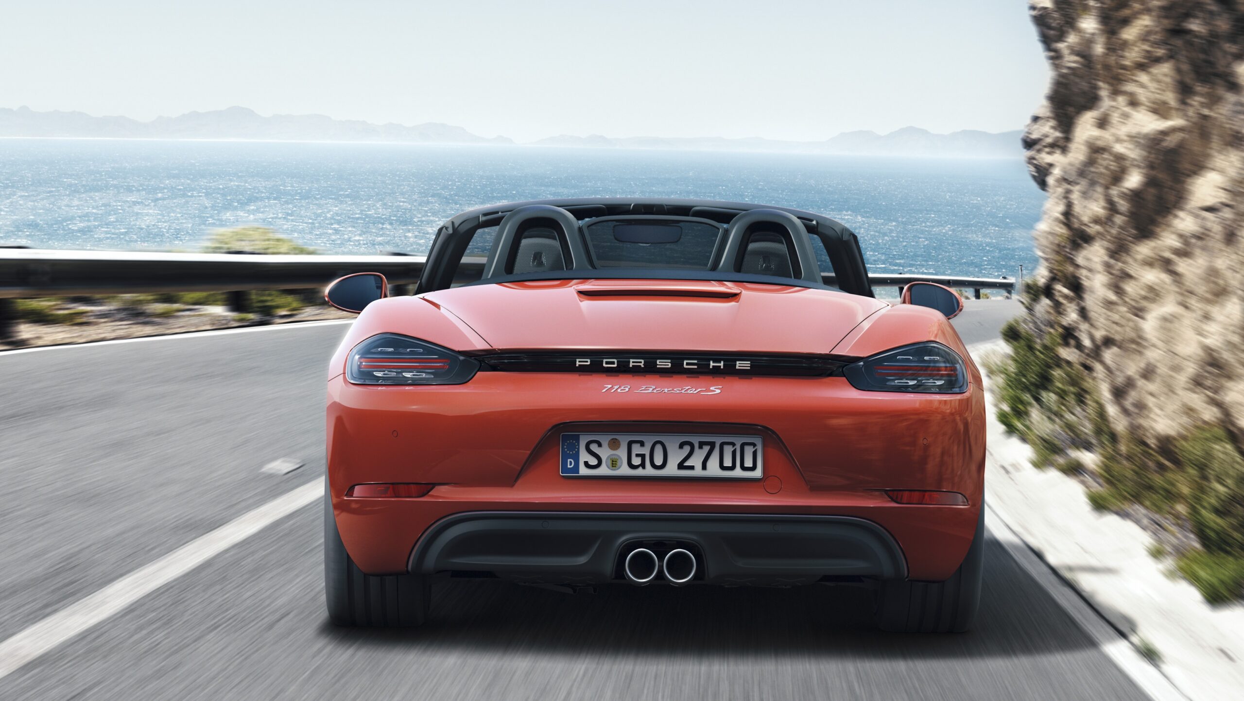 Official Porsche photo of Lava Orange 718 Boxster S rear open-top on coastal road