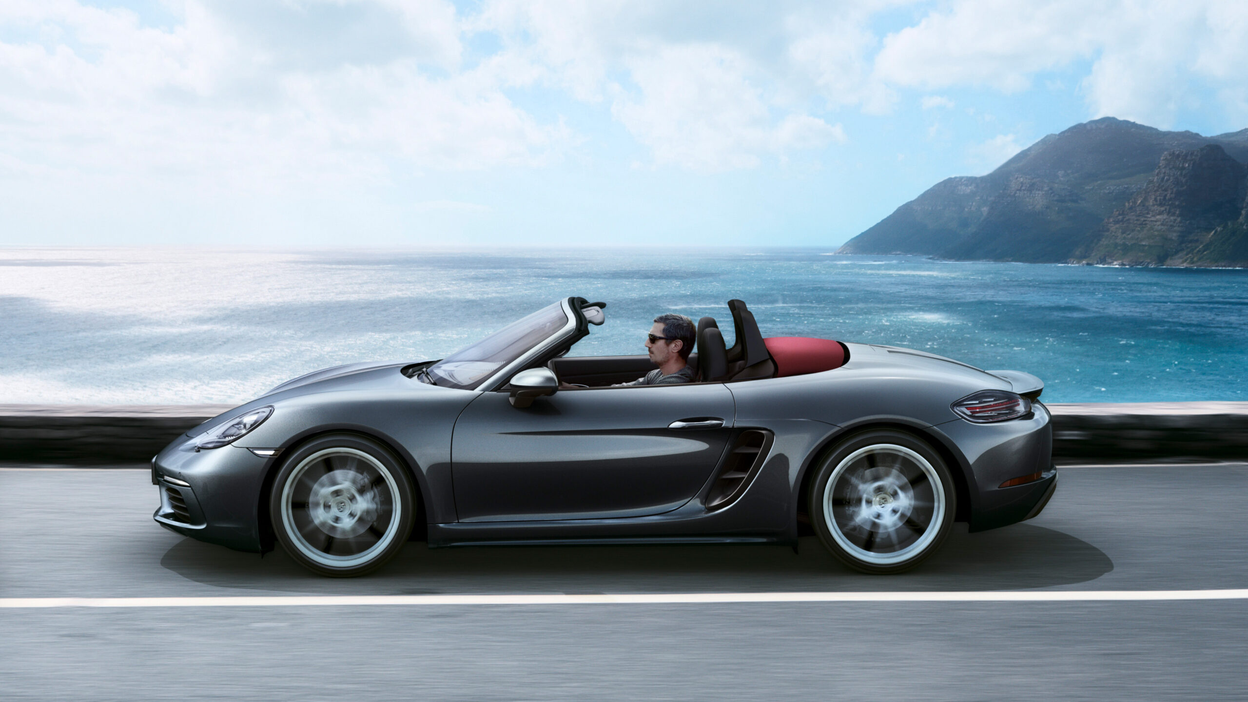 Official Porsche photo of Agate Grey 718 Boxster side profile soft-top down on coastal highway