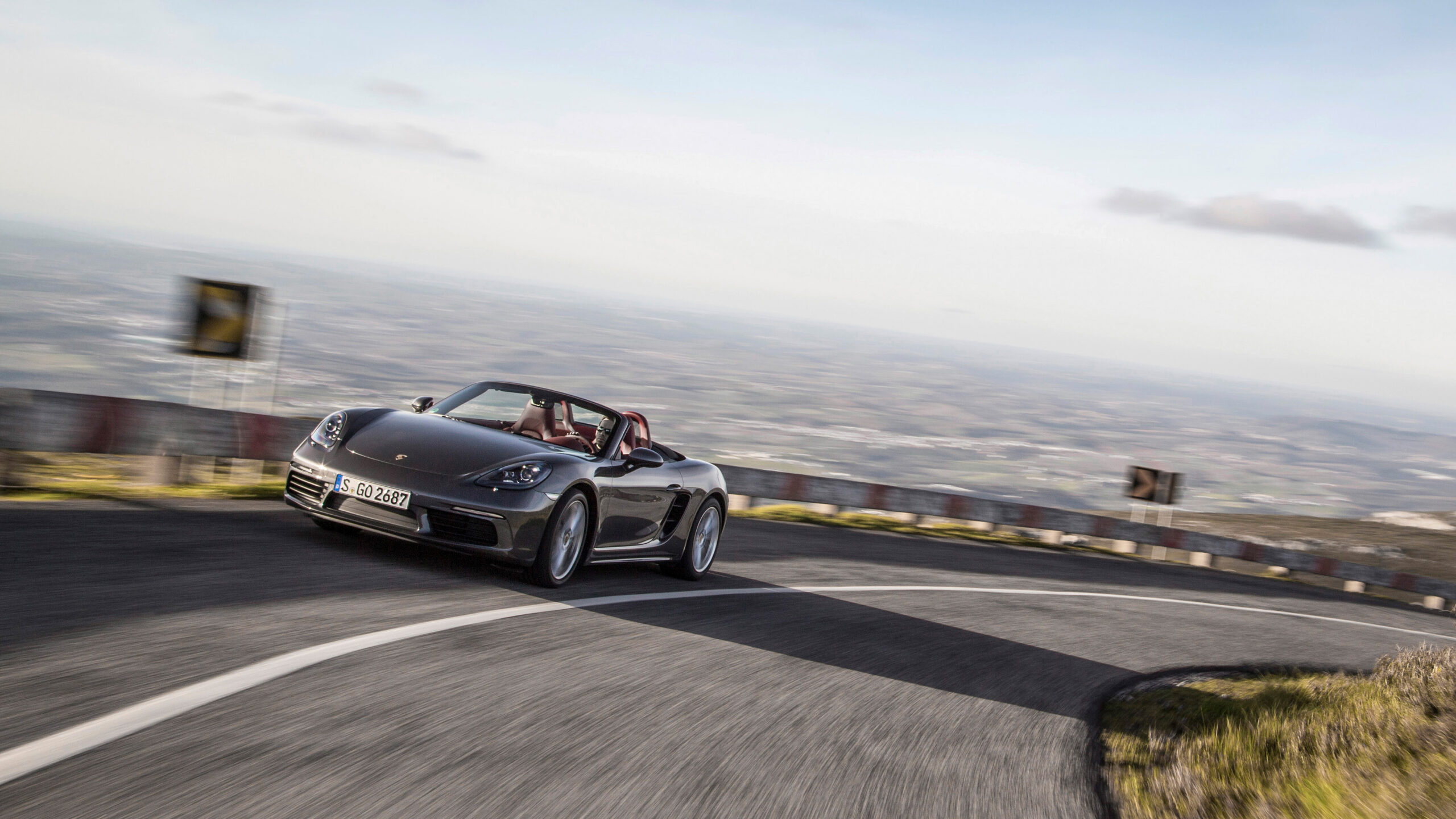 Official Porsche photo of Agate Grey 718 Boxster cornering open-top on mountain road