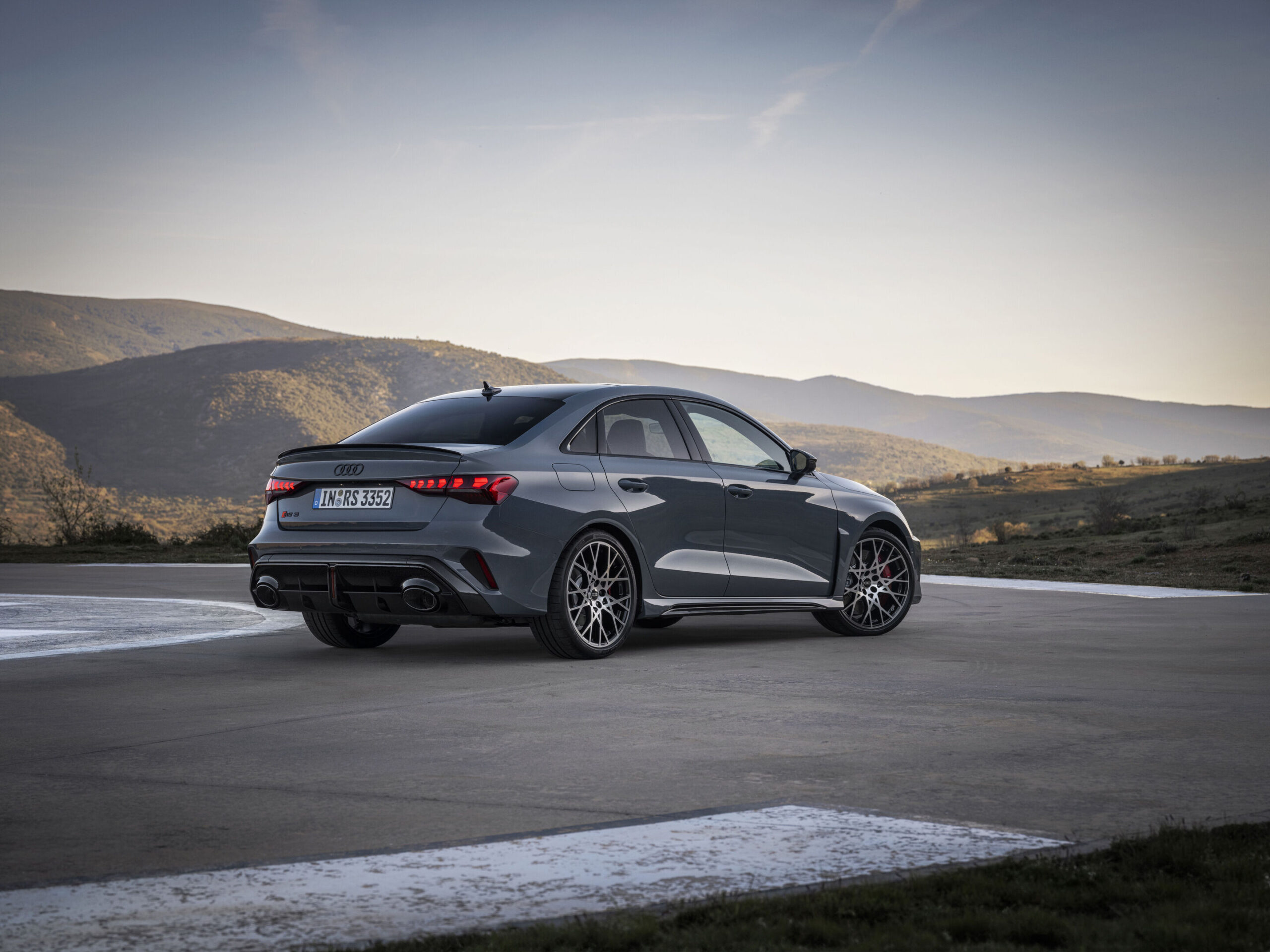 Official Audi photo of a Kemora Gray 2026 Audi RS 3 Sedan rear three-quarter on a pad in the hills