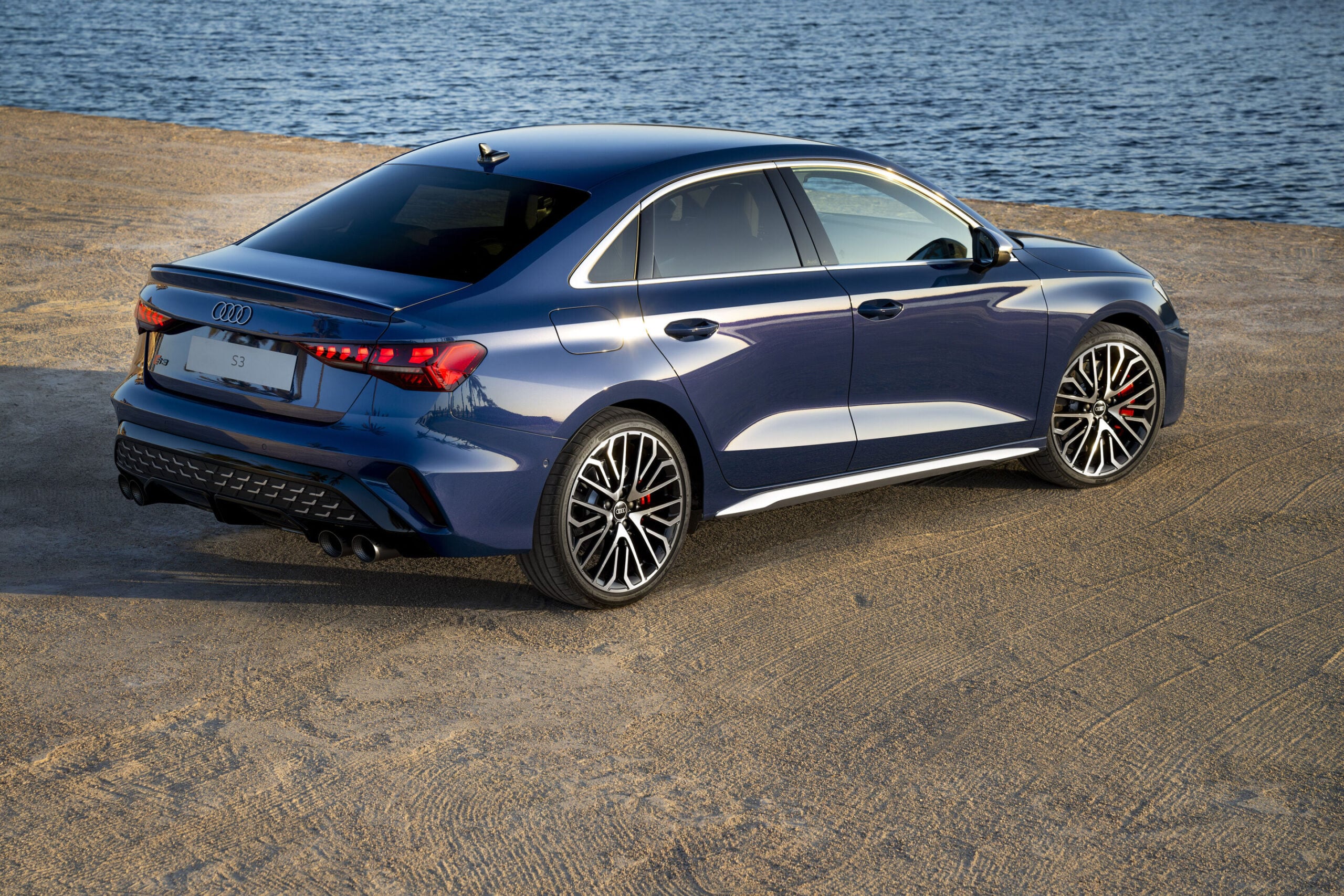 Official Audi photo of a dark blue 2026 Audi S3 rear three-quarter with quad exhausts by water
